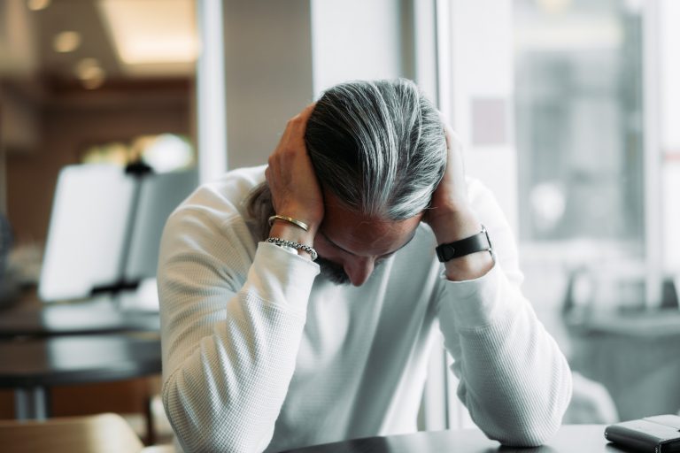 Person with gray hair sitting at a desk, hands on their head in a stressed pose in an office setting.