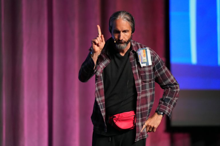 Male performer on stage with headset mic, raising his index finger, wearing a plaid shirt and a red waist pouch.