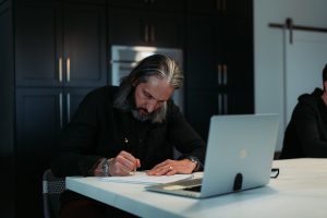 Man with gray hair and beard writing in a notebook at a white table, with a laptop nearby in a dark kitchen setting.