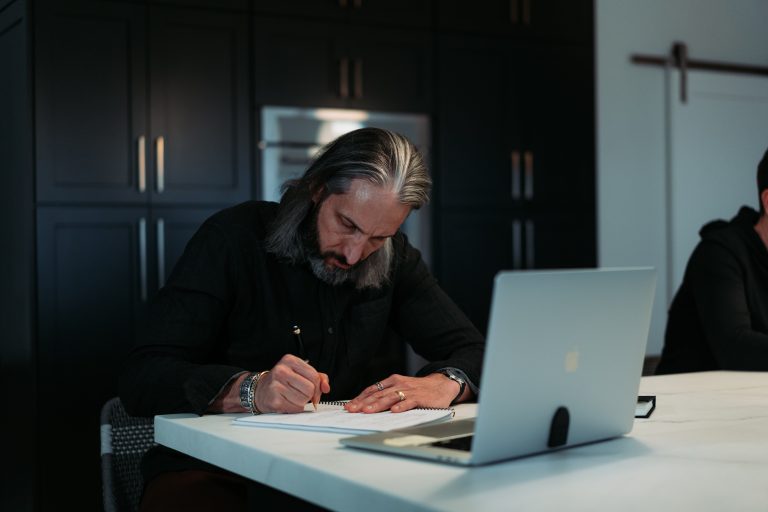 Man with gray hair and beard writing in a notebook at a white table, with a laptop nearby in a dark kitchen setting.
