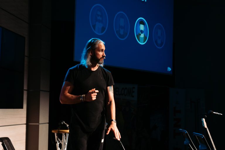 Male speaker on stage in black outfit wearing a headset mic, gesturing with his hand against a dark background and blue screen behind him.