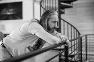 Bearded man with long hair rests his arms on a metal railing, gazing downward in an outdoor setting (black and white).