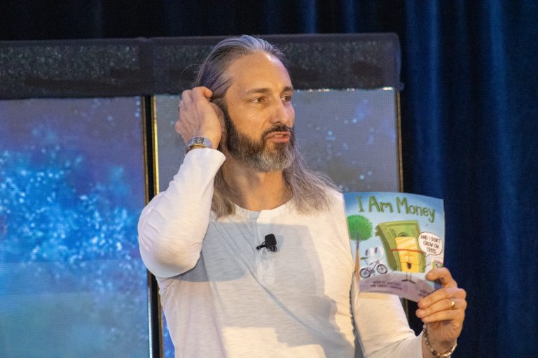 Bearded man with long hair in a light shirt on stage, reading a children’s book titled 'I Am Money'.