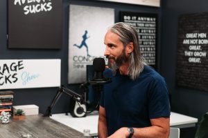 Man with long gray hair and beard sits at a podcast microphone in a recording studio, speaking into the mic with posters on dark walls in the background.